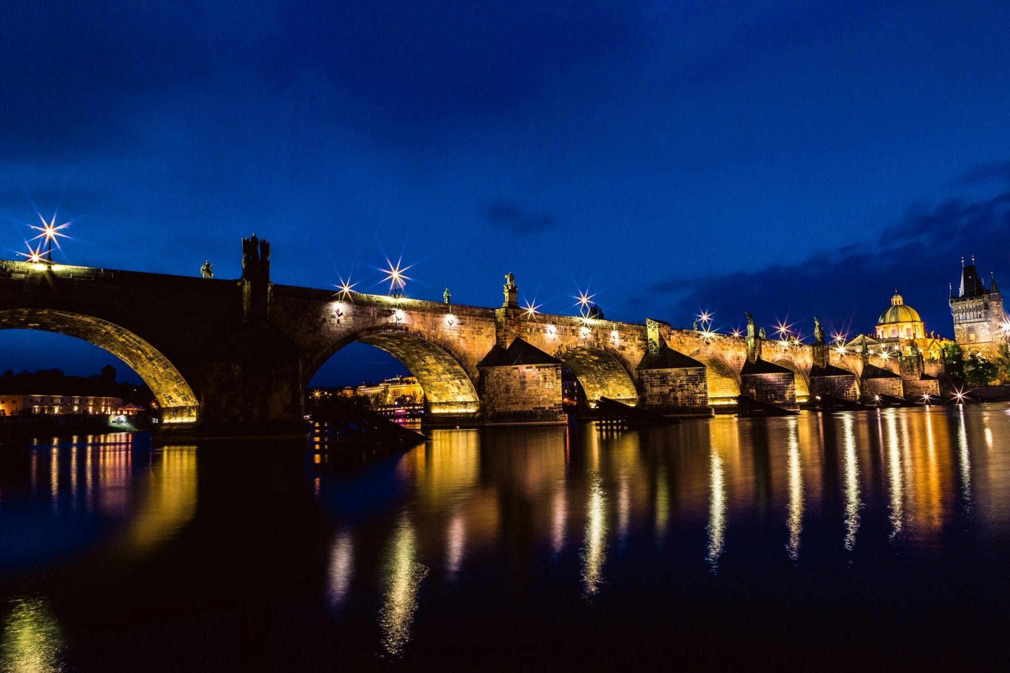 Praga al anochecer: faroles antiguos junto al río Moldava y el Puente de Carlos iluminado suavemente.